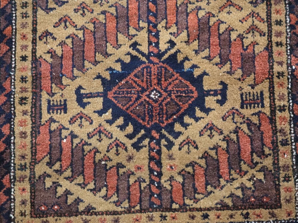 An antique Baluch hand-knotted wool rug featuring three diamond latch-hook medallions on a camel-coloured field with geometric red and navy borders. View XII