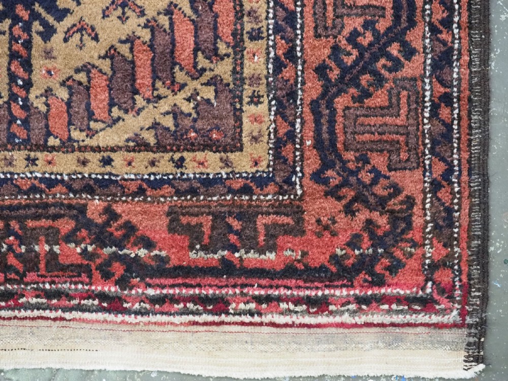 An antique Baluch hand-knotted wool rug featuring three diamond latch-hook medallions on a camel-coloured field with geometric red and navy borders. View XIII