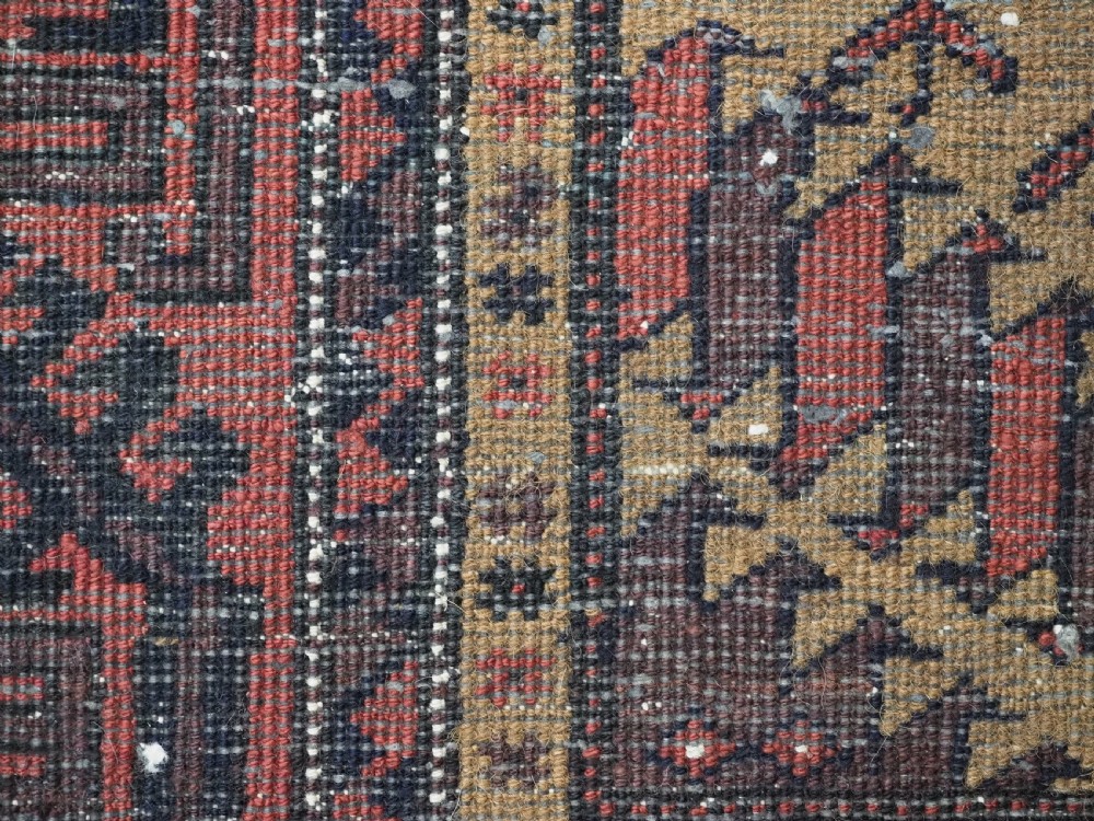 An antique Baluch hand-knotted wool rug featuring three diamond latch-hook medallions on a camel-coloured field with geometric red and navy borders. View XIV