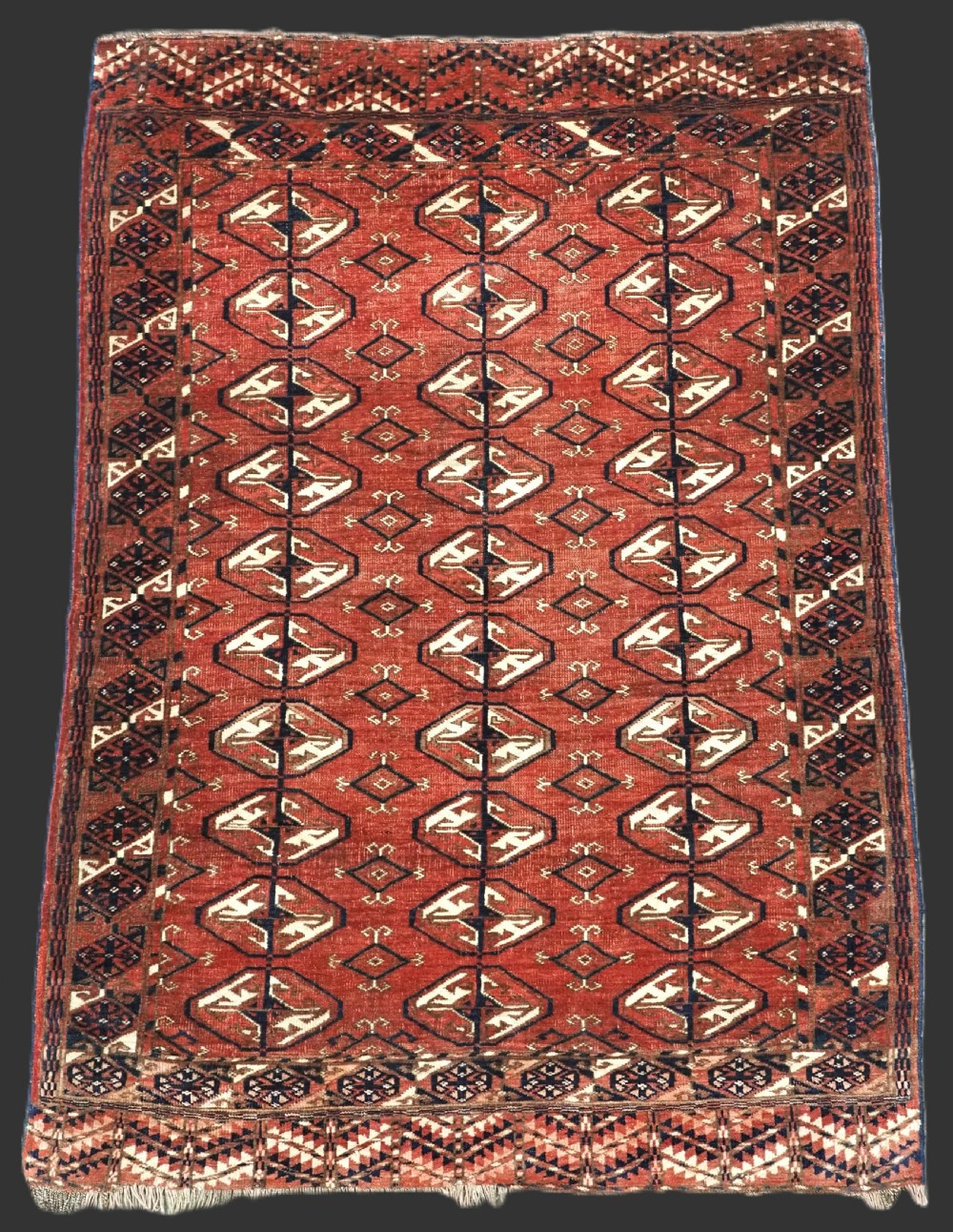 A hand-knotted antique Tekke Turkmen wool rug featuring repeating ovoid gul motifs set against a deep terracotta red background with geometric borders.