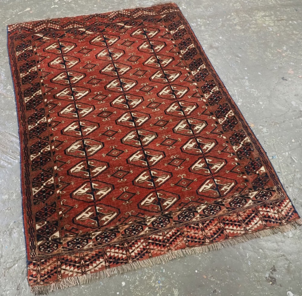 A hand-knotted antique Tekke Turkmen wool rug featuring repeating ovoid gul motifs set against a deep terracotta red background with geometric borders. View II