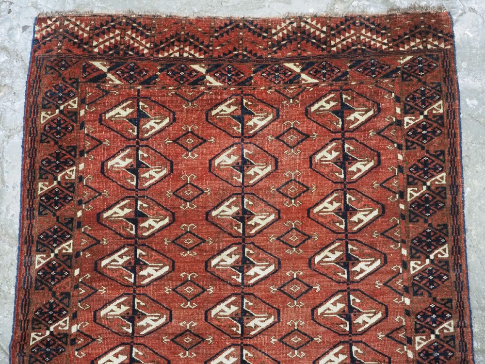 A hand-knotted antique Tekke Turkmen wool rug featuring repeating ovoid gul motifs set against a deep terracotta red background with geometric borders. View III