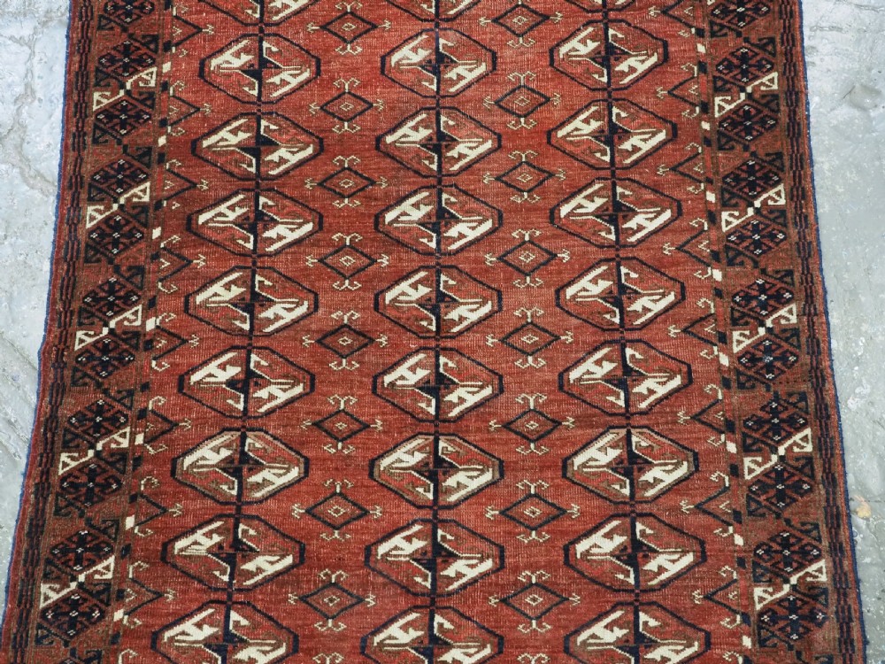 A hand-knotted antique Tekke Turkmen wool rug featuring repeating ovoid gul motifs set against a deep terracotta red background with geometric borders. View IV