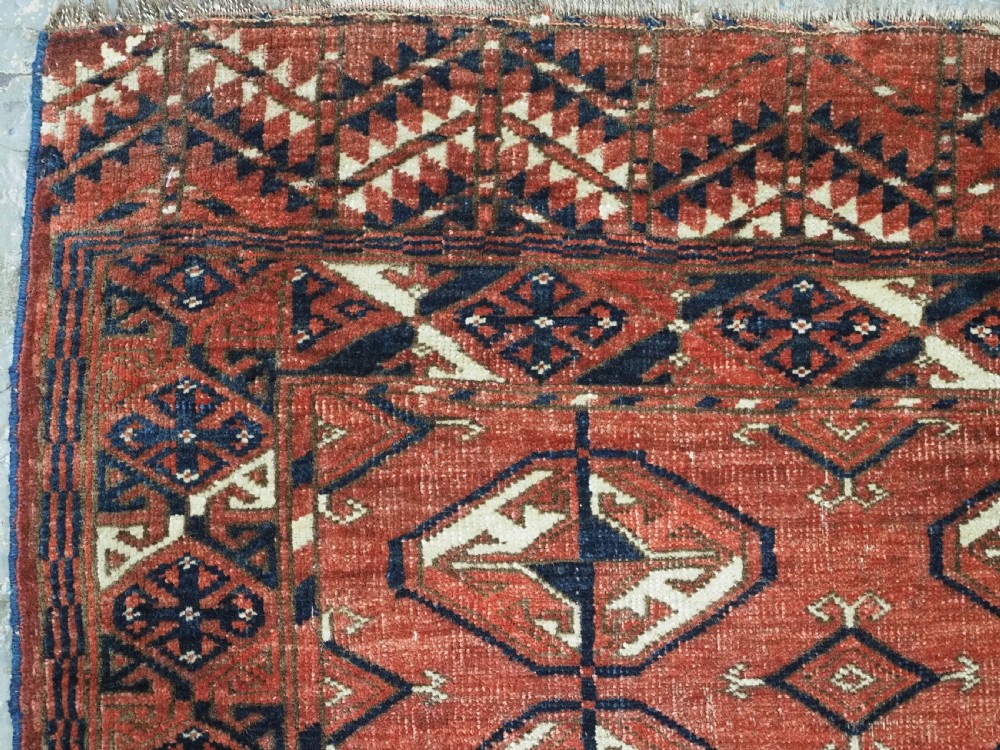 A hand-knotted antique Tekke Turkmen wool rug featuring repeating ovoid gul motifs set against a deep terracotta red background with geometric borders. View V