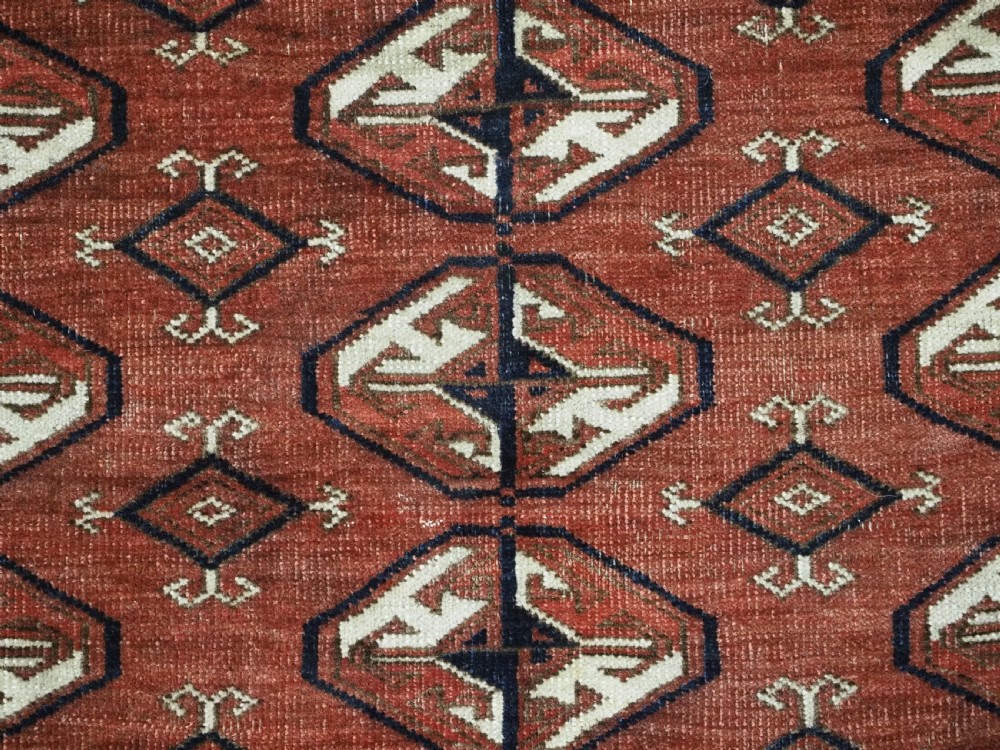 A hand-knotted antique Tekke Turkmen wool rug featuring repeating ovoid gul motifs set against a deep terracotta red background with geometric borders. View IX