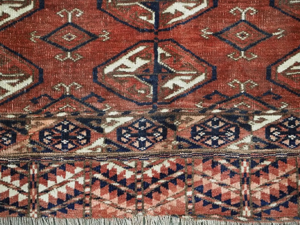 A hand-knotted antique Tekke Turkmen wool rug featuring repeating ovoid gul motifs set against a deep terracotta red background with geometric borders. View X