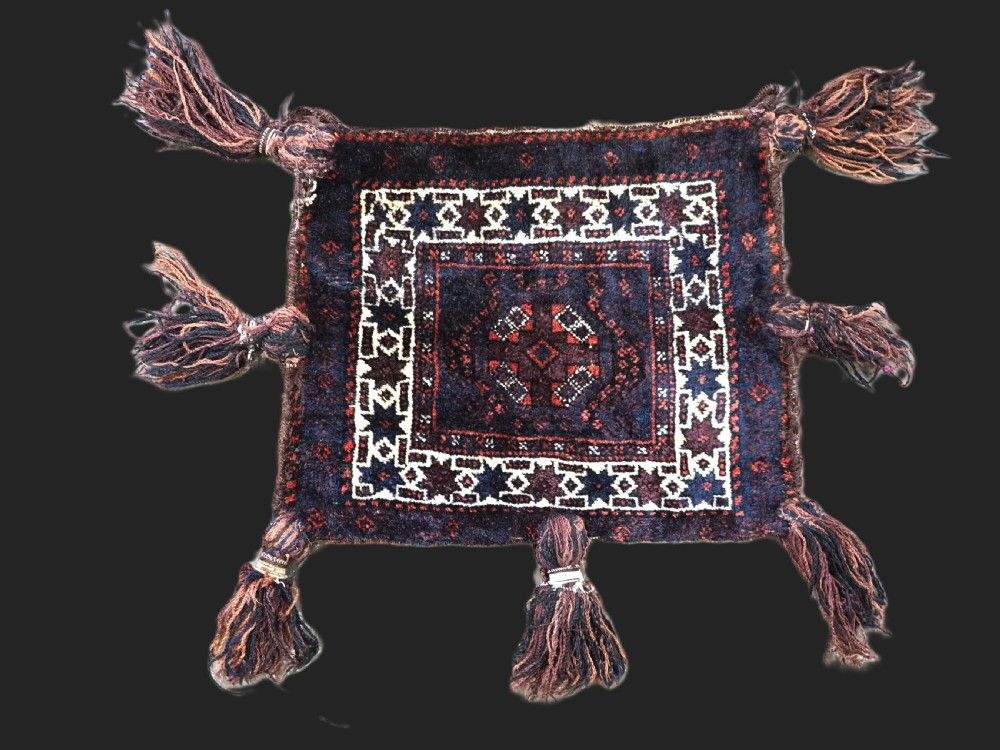 A square dark navy blue and terracotta red handwoven Baluch tribal vanity bag featuring geometric central motifs, white borders, and prominent wool tassels along the edges.