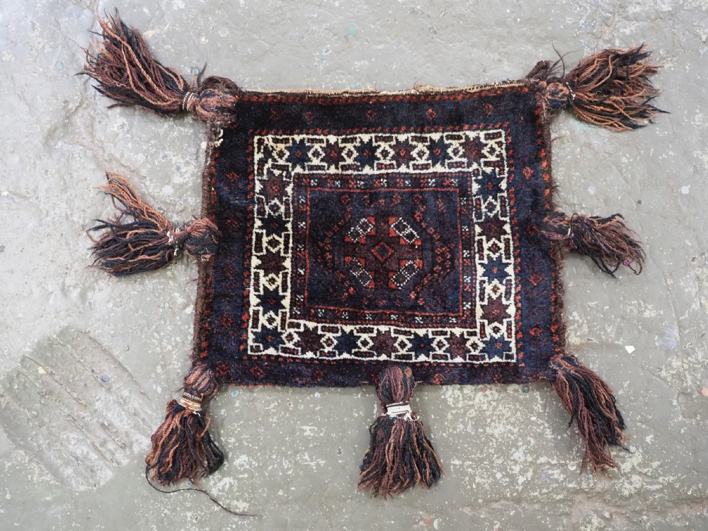 A square dark navy blue and terracotta red handwoven Baluch tribal vanity bag featuring geometric central motifs, white borders, and prominent wool tassels along the edges. View II