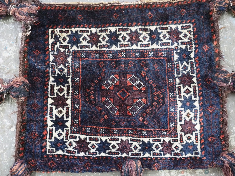A square dark navy blue and terracotta red handwoven Baluch tribal vanity bag featuring geometric central motifs, white borders, and prominent wool tassels along the edges. View III