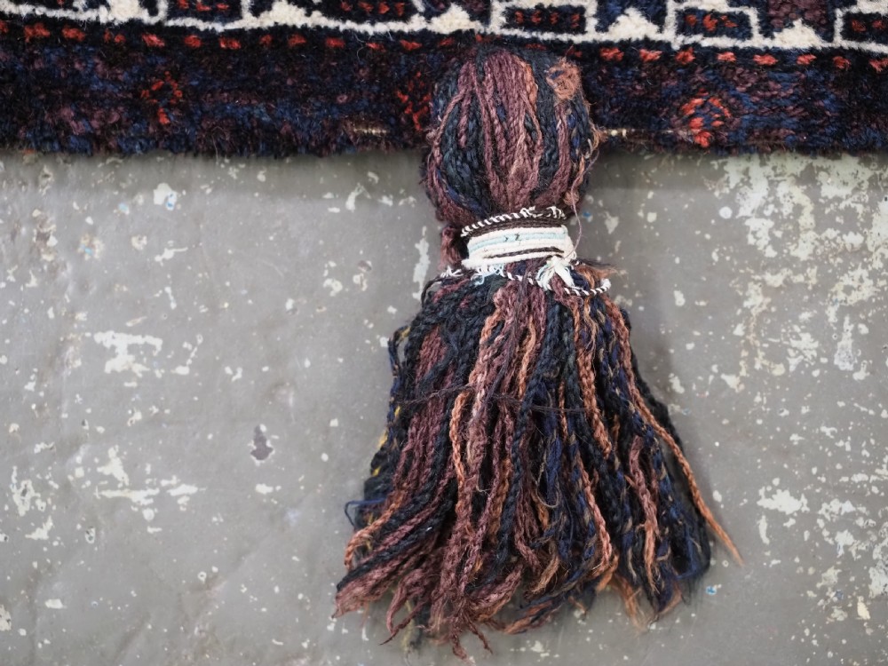 A square dark navy blue and terracotta red handwoven Baluch tribal vanity bag featuring geometric central motifs, white borders, and prominent wool tassels along the edges. View V