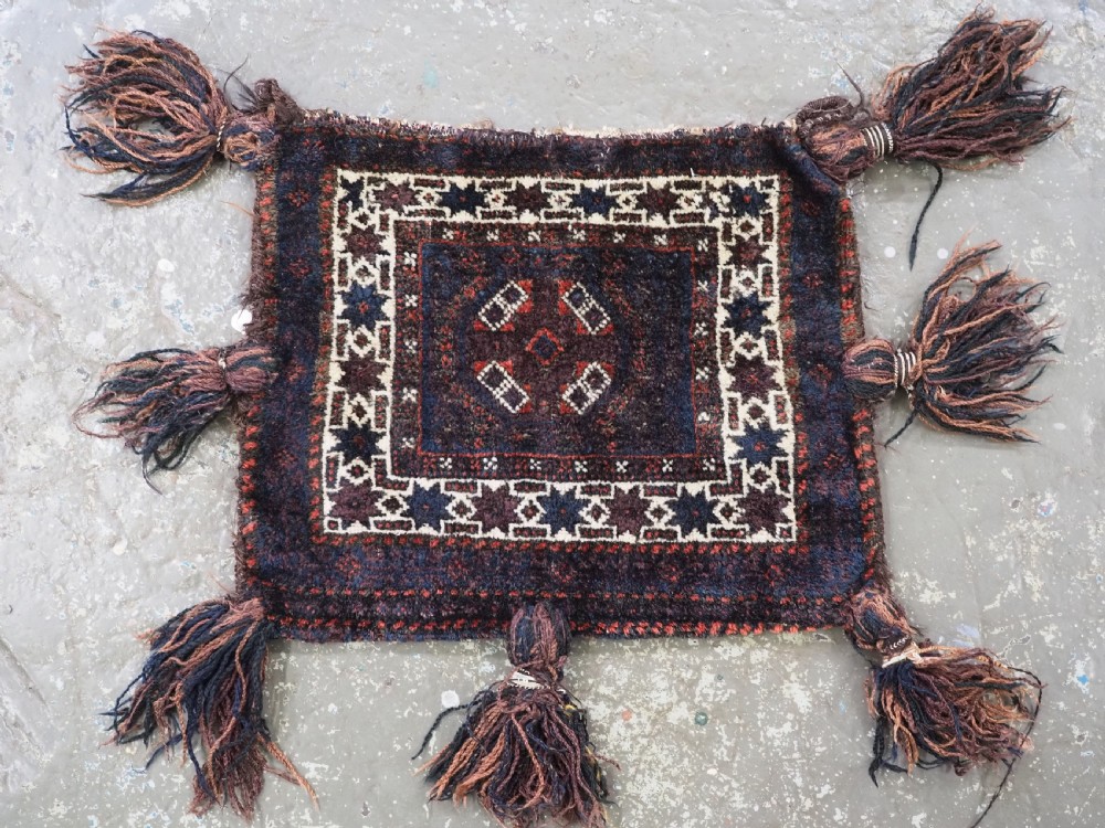A square dark navy blue and terracotta red handwoven Baluch tribal vanity bag featuring geometric central motifs, white borders, and prominent wool tassels along the edges. View VI