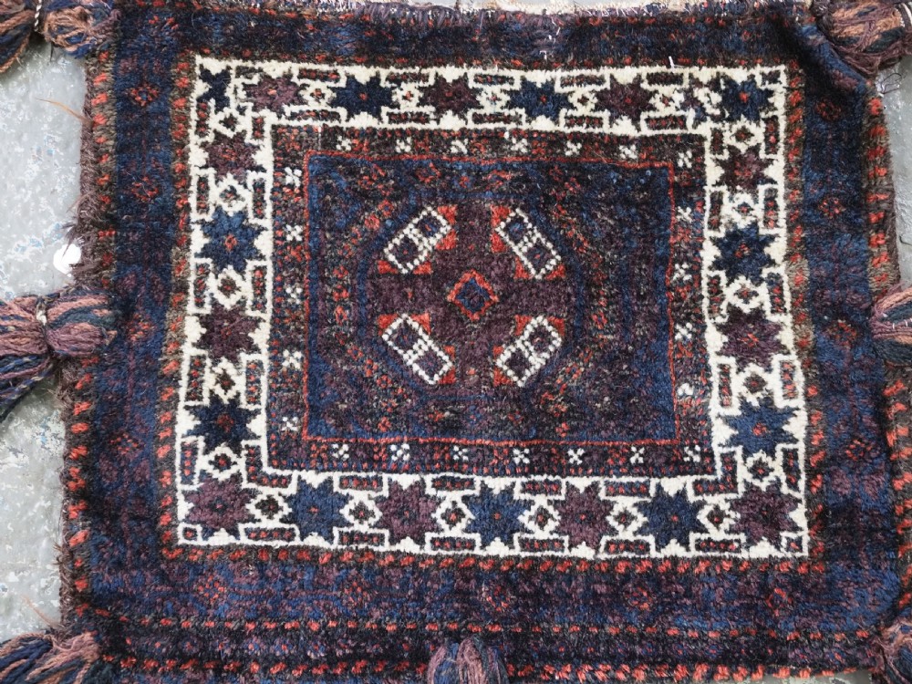 A square dark navy blue and terracotta red handwoven Baluch tribal vanity bag featuring geometric central motifs, white borders, and prominent wool tassels along the edges. View VII