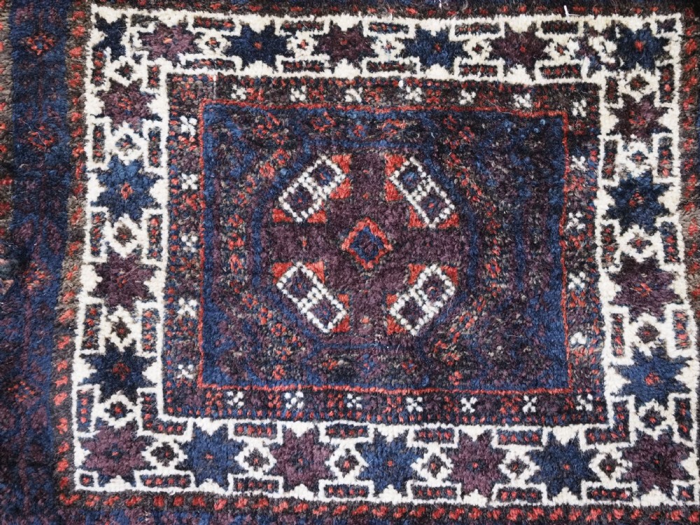 A square dark navy blue and terracotta red handwoven Baluch tribal vanity bag featuring geometric central motifs, white borders, and prominent wool tassels along the edges. View VIII