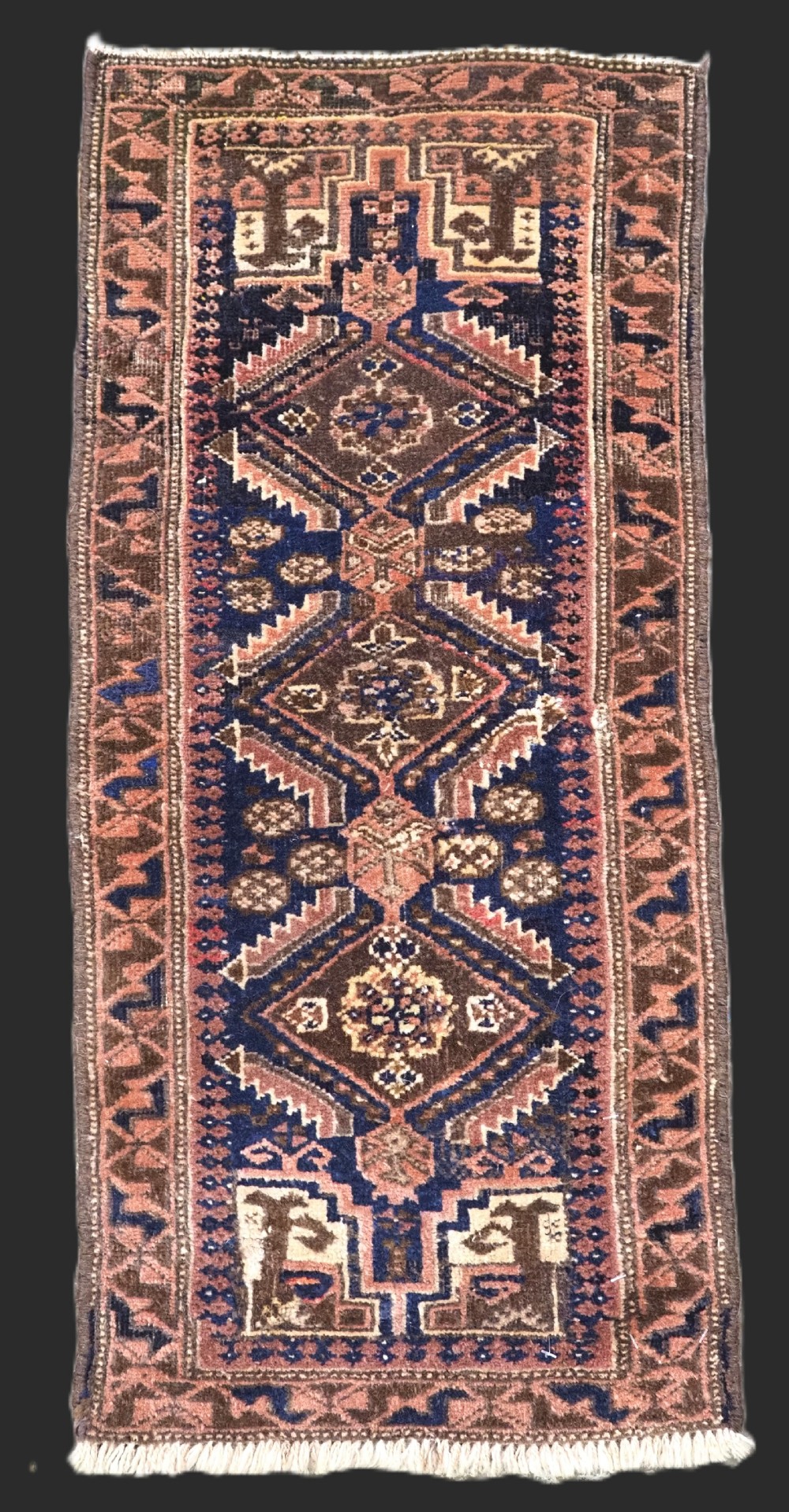 A small hand-knotted rectangular Baluch tribal rug face featuring geometric diamond medallions in indigo blue, terracotta, and brown tones with fringed ends.