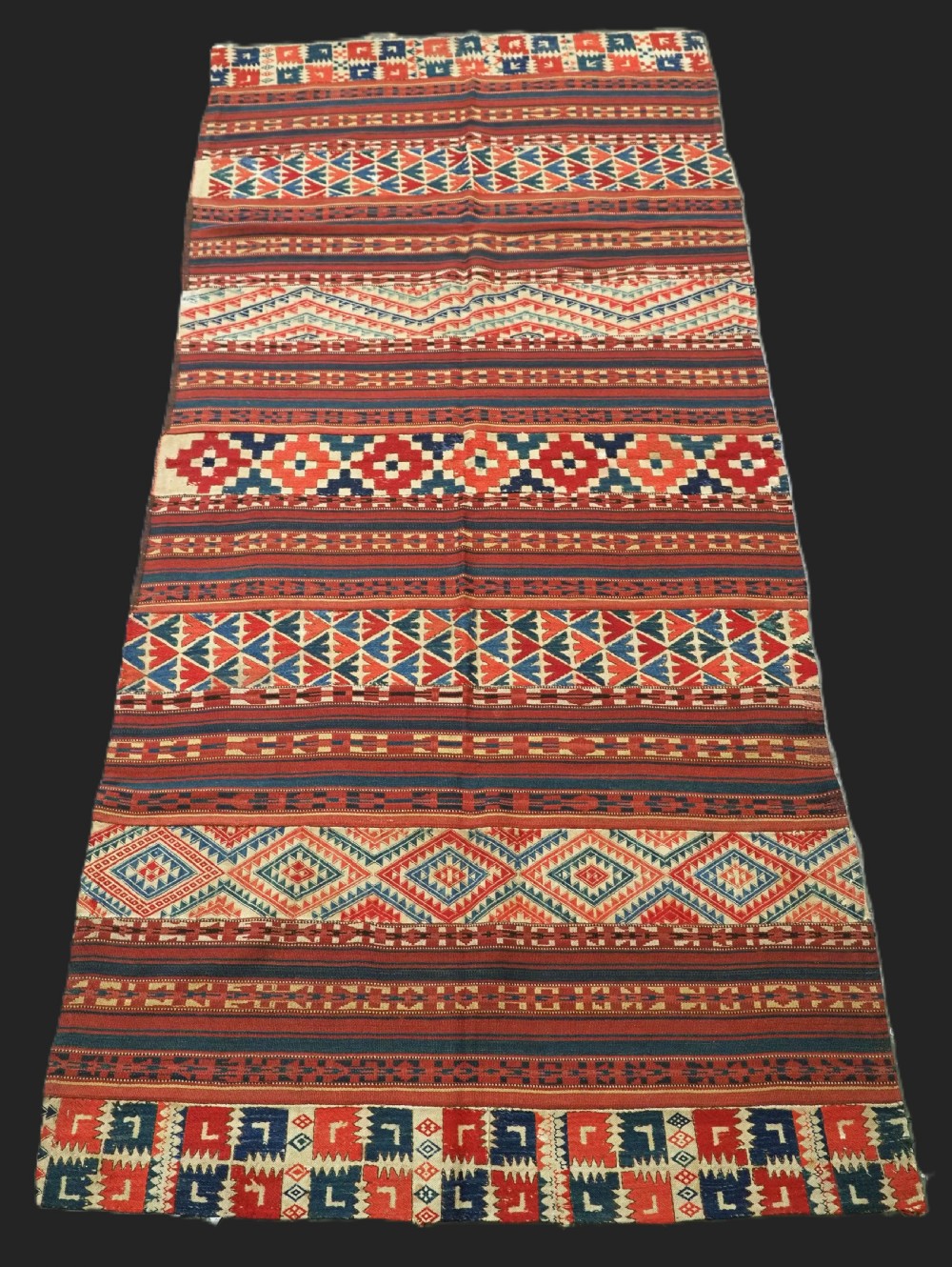 An antique central Asian kilim rug composed of thirteen vertical strips showing geometric diamond and stripe patterns in red, navy blue, and cream.