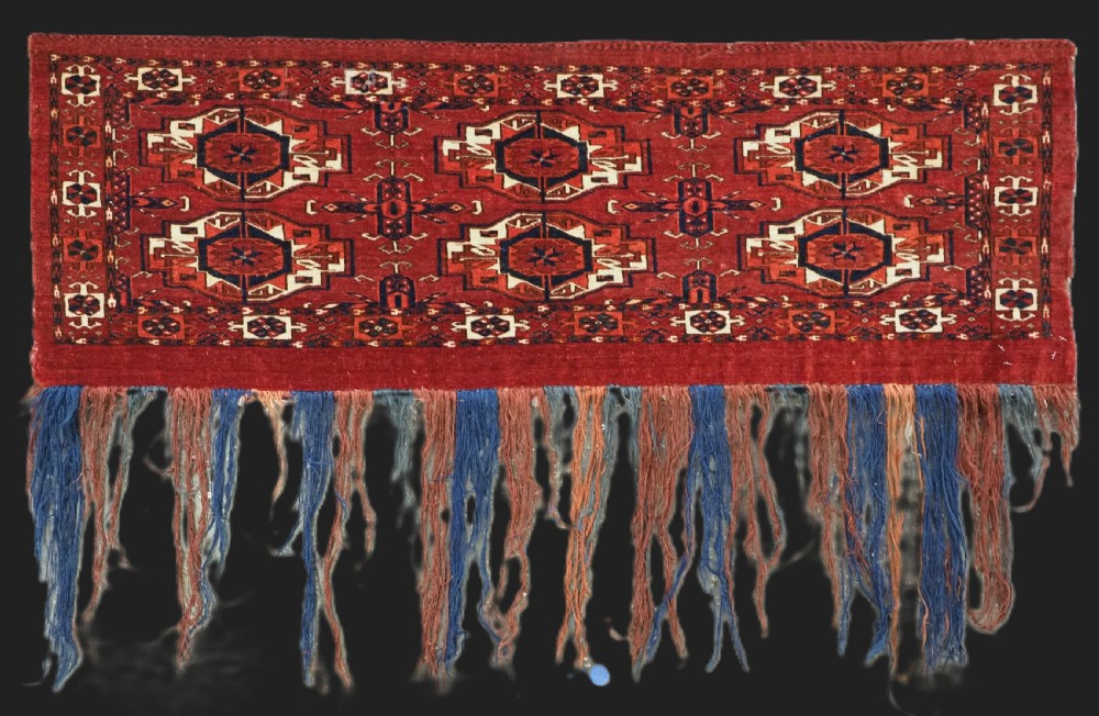 A rectangular hand-woven Tekke Turkmen wool textile featuring six octagonal guls on a deep red field with an intricate geometric border and long blue and red fringe along the bottom edge.