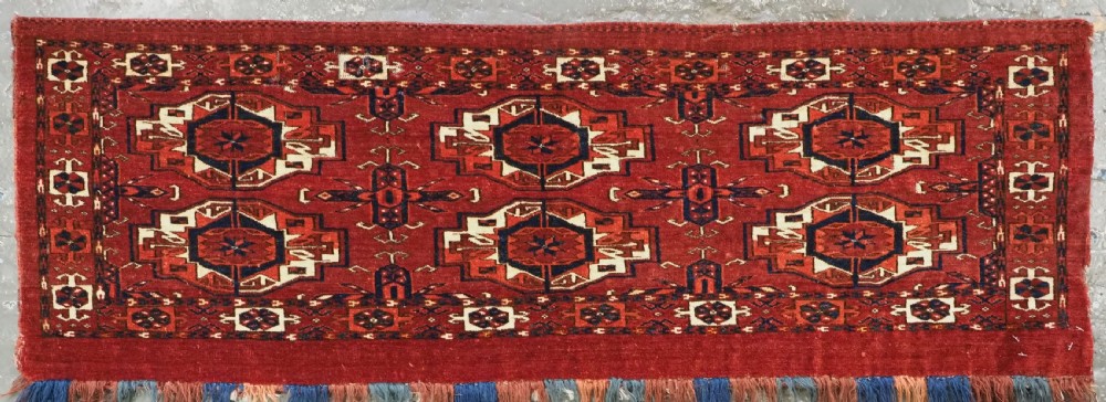 A rectangular hand-woven Tekke Turkmen wool textile featuring six octagonal guls on a deep red field with an intricate geometric border and long blue and red fringe along the bottom edge. View III