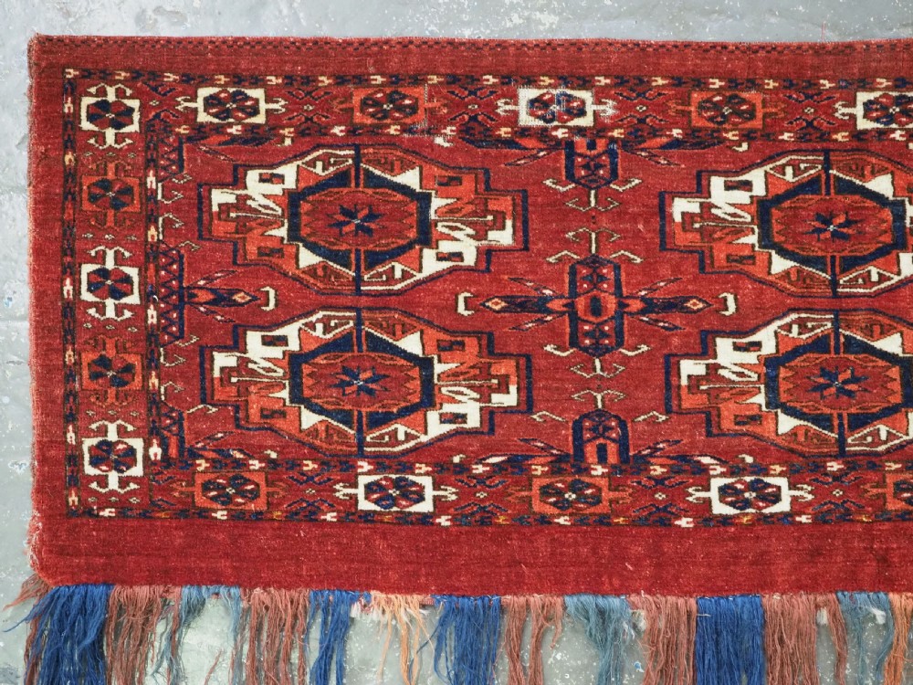 A rectangular hand-woven Tekke Turkmen wool textile featuring six octagonal guls on a deep red field with an intricate geometric border and long blue and red fringe along the bottom edge. View IV
