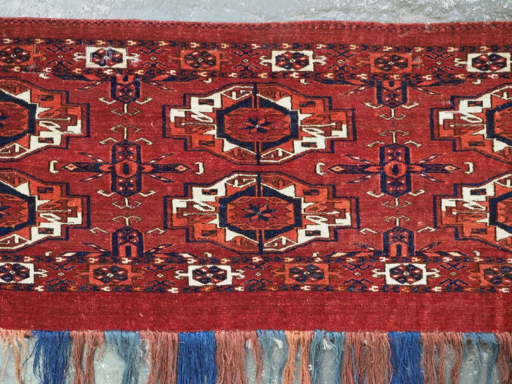 A rectangular hand-woven Tekke Turkmen wool textile featuring six octagonal guls on a deep red field with an intricate geometric border and long blue and red fringe along the bottom edge. View V