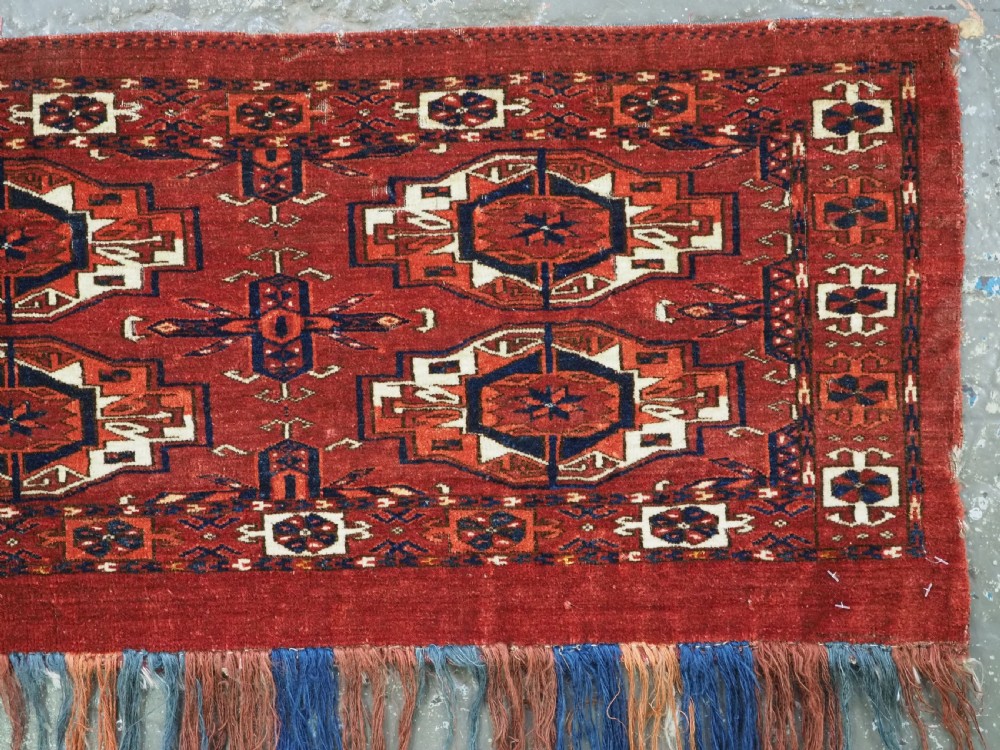 A rectangular hand-woven Tekke Turkmen wool textile featuring six octagonal guls on a deep red field with an intricate geometric border and long blue and red fringe along the bottom edge. View VI
