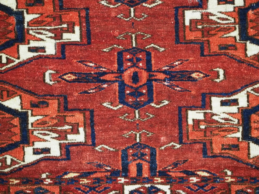 A rectangular hand-woven Tekke Turkmen wool textile featuring six octagonal guls on a deep red field with an intricate geometric border and long blue and red fringe along the bottom edge. View X