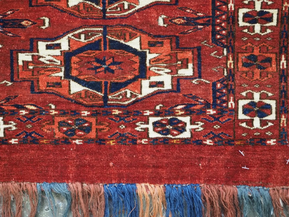 A rectangular hand-woven Tekke Turkmen wool textile featuring six octagonal guls on a deep red field with an intricate geometric border and long blue and red fringe along the bottom edge. View XI