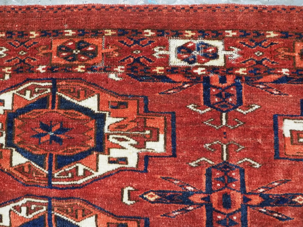 A rectangular hand-woven Tekke Turkmen wool textile featuring six octagonal guls on a deep red field with an intricate geometric border and long blue and red fringe along the bottom edge. View XIII
