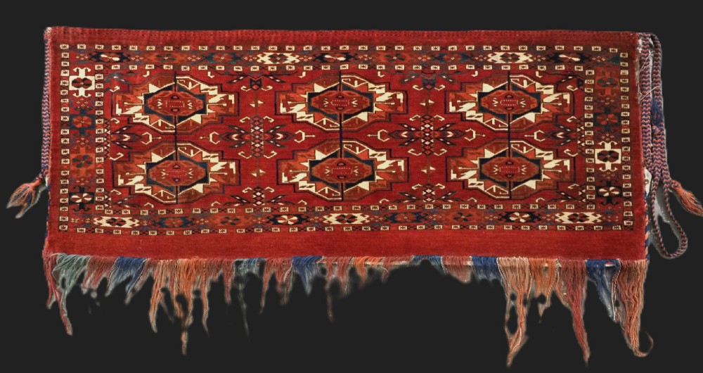 A horizontal handwoven wool Tekke Turkmen torba featuring six octagonal gul medallions on a deep red background with geometric borders and braided side tassels.