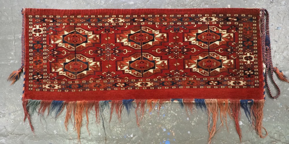 A horizontal handwoven wool Tekke Turkmen torba featuring six octagonal gul medallions on a deep red background with geometric borders and braided side tassels. View II