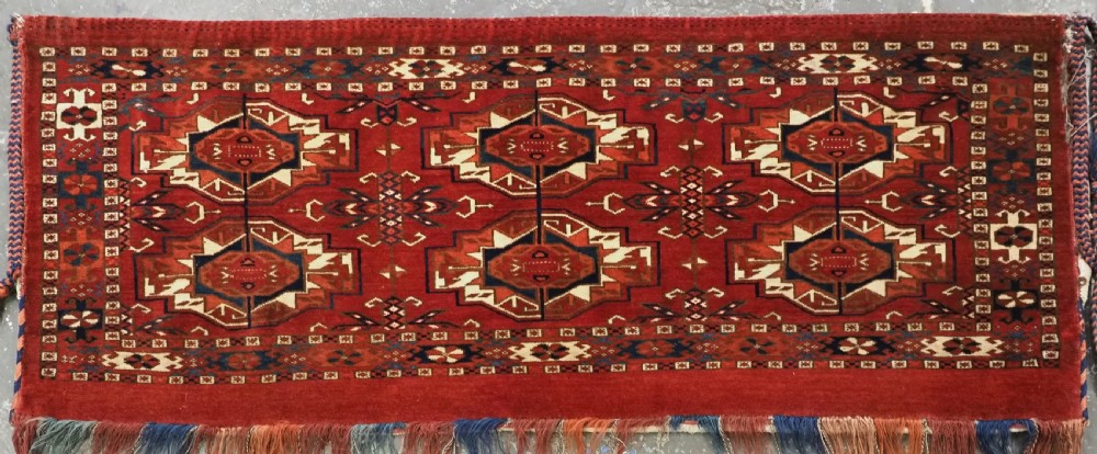 A horizontal handwoven wool Tekke Turkmen torba featuring six octagonal gul medallions on a deep red background with geometric borders and braided side tassels. View III