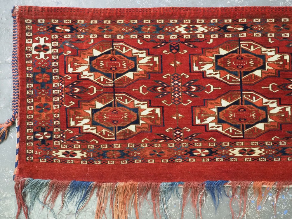 A horizontal handwoven wool Tekke Turkmen torba featuring six octagonal gul medallions on a deep red background with geometric borders and braided side tassels. View IV