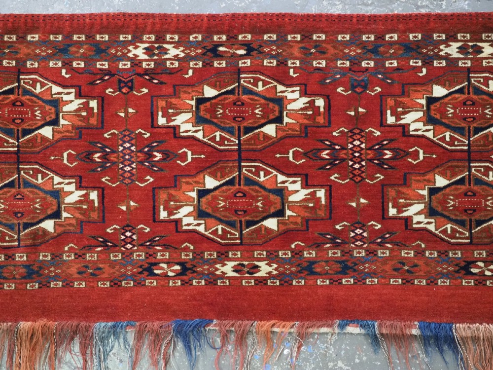 A horizontal handwoven wool Tekke Turkmen torba featuring six octagonal gul medallions on a deep red background with geometric borders and braided side tassels. View V