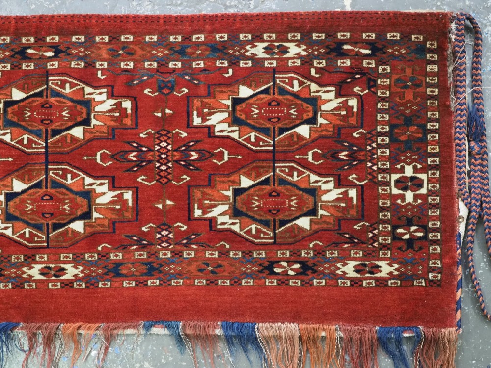 A horizontal handwoven wool Tekke Turkmen torba featuring six octagonal gul medallions on a deep red background with geometric borders and braided side tassels. View VI