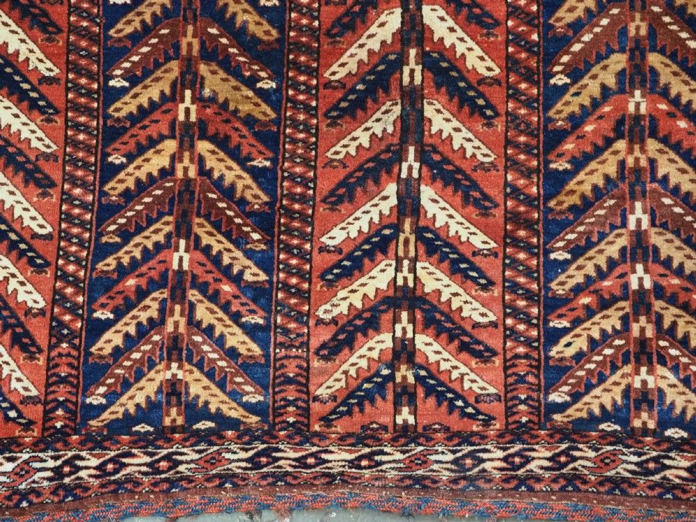 A pentagonal antique Turkmen wool textile featuring vertical rows of white and gold chevron leaf motifs on a dark red background framed by geometric borders. View VI