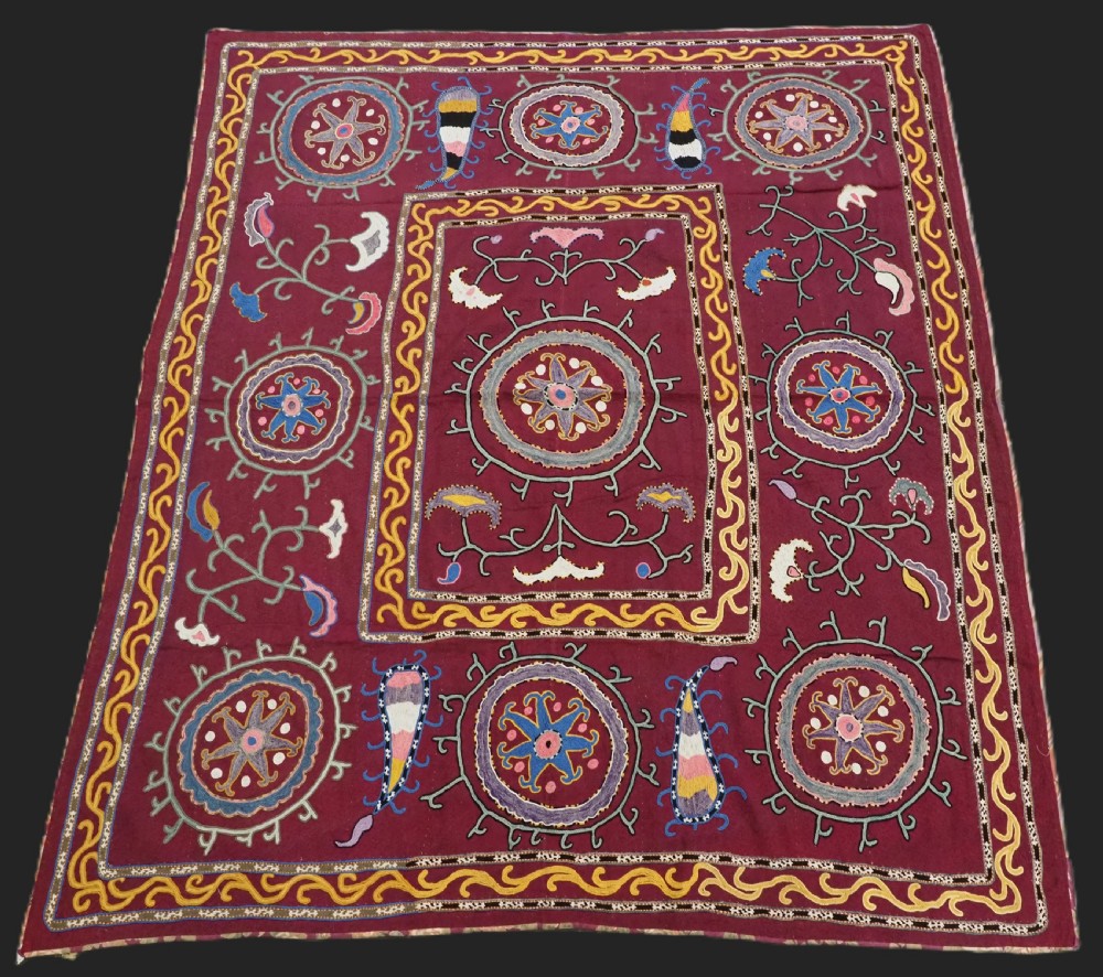 A rectangular antique crimson red textile featuring intricate silk embroidery with solar medallion motifs, floral scrolls, and geometric borders in yellow, blue, and pink.