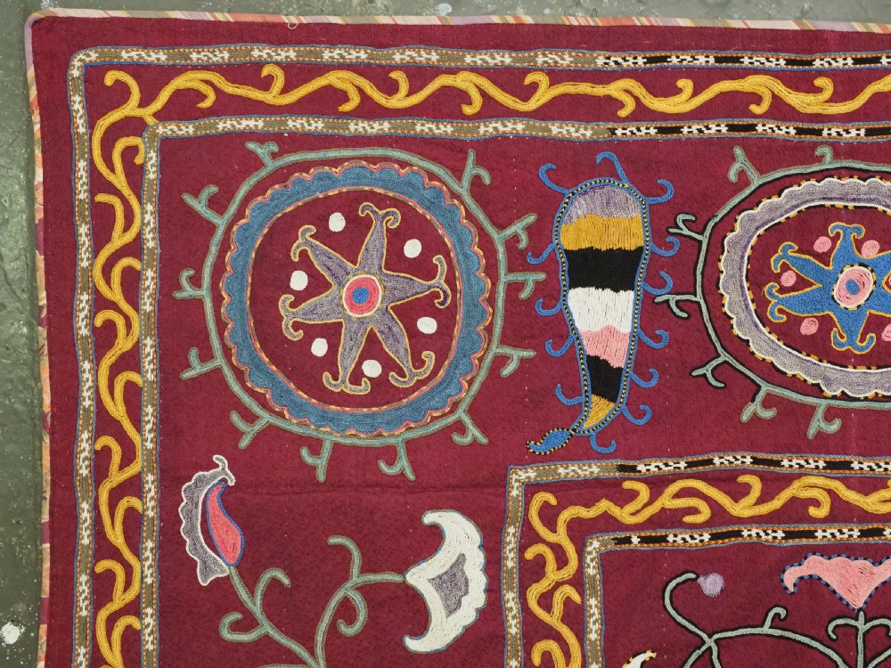 A rectangular antique crimson red textile featuring intricate silk embroidery with solar medallion motifs, floral scrolls, and geometric borders in yellow, blue, and pink. View II