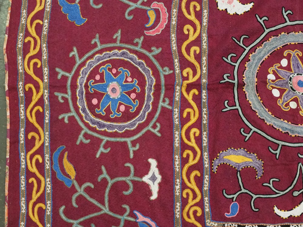 A rectangular antique crimson red textile featuring intricate silk embroidery with solar medallion motifs, floral scrolls, and geometric borders in yellow, blue, and pink. View III