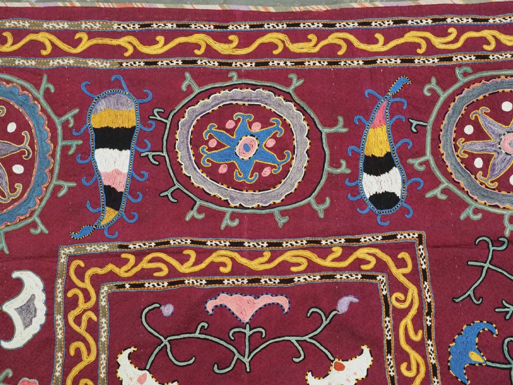 A rectangular antique crimson red textile featuring intricate silk embroidery with solar medallion motifs, floral scrolls, and geometric borders in yellow, blue, and pink. View IV
