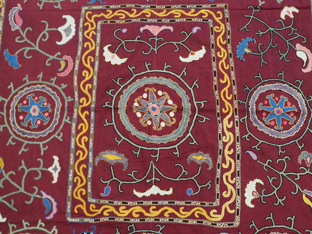 A rectangular antique crimson red textile featuring intricate silk embroidery with solar medallion motifs, floral scrolls, and geometric borders in yellow, blue, and pink. View V