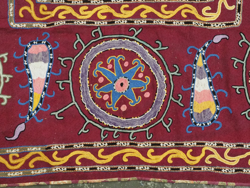 A rectangular antique crimson red textile featuring intricate silk embroidery with solar medallion motifs, floral scrolls, and geometric borders in yellow, blue, and pink. View VI