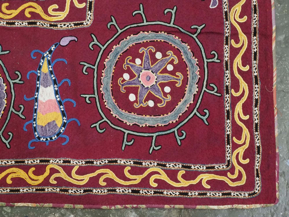 A rectangular antique crimson red textile featuring intricate silk embroidery with solar medallion motifs, floral scrolls, and geometric borders in yellow, blue, and pink. View VII