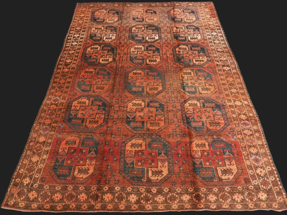 An antique northern Afghan Ersari Turkmen wool carpet featuring repeating octagonal gul motifs in dark blue, beige, and brown set against a deep terracotta red field.
