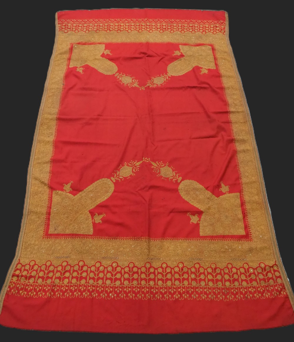 A rectangular red wool shawl with extensive gold-coloured needlework along the borders and corner motifs.
