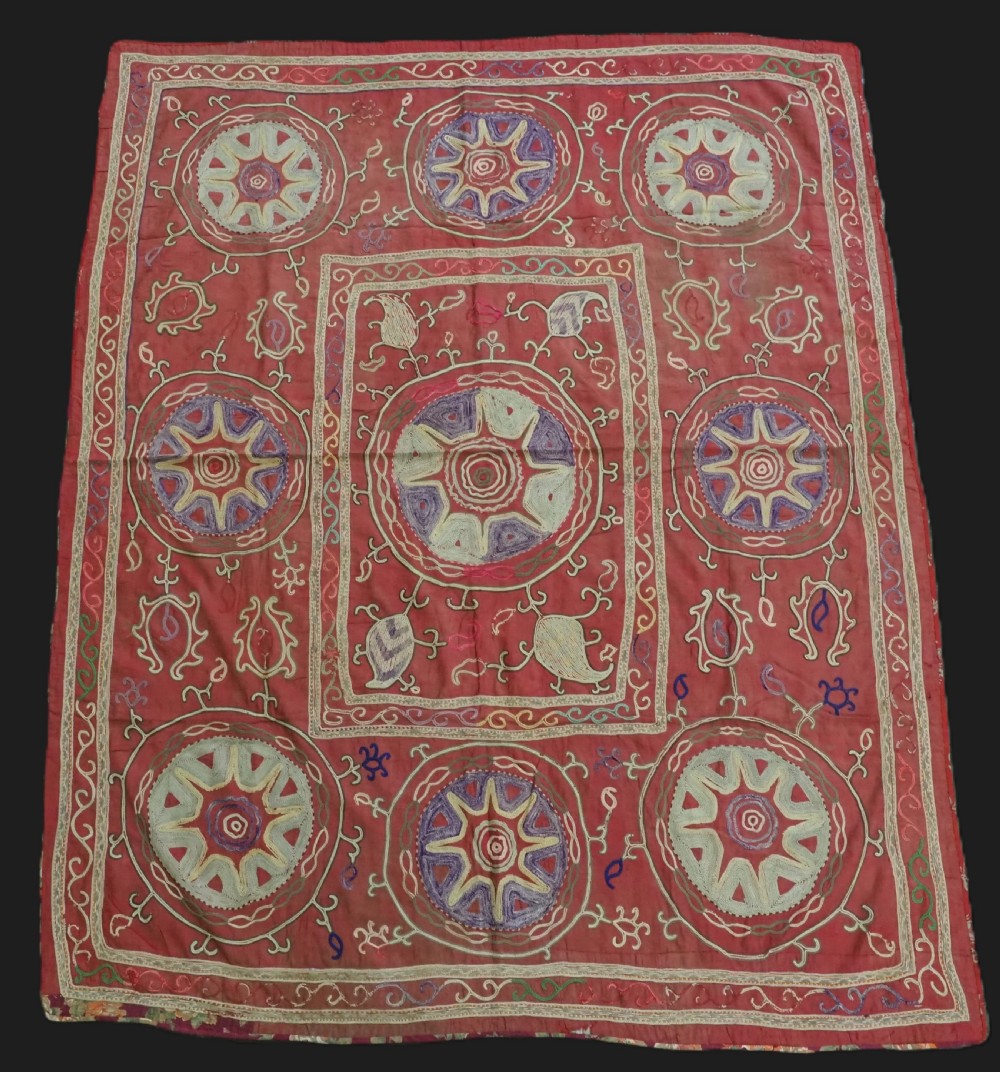 A rectangular red cotton embroidered suzani featuring geometric starburst medallions and floral scrolling borders worked in multi-coloured silk thread.