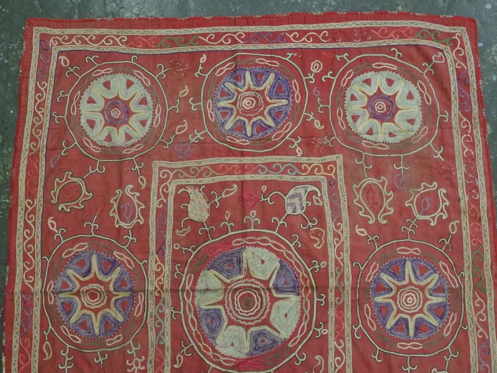 A rectangular red cotton embroidered suzani featuring geometric starburst medallions and floral scrolling borders worked in multi-coloured silk thread. View II