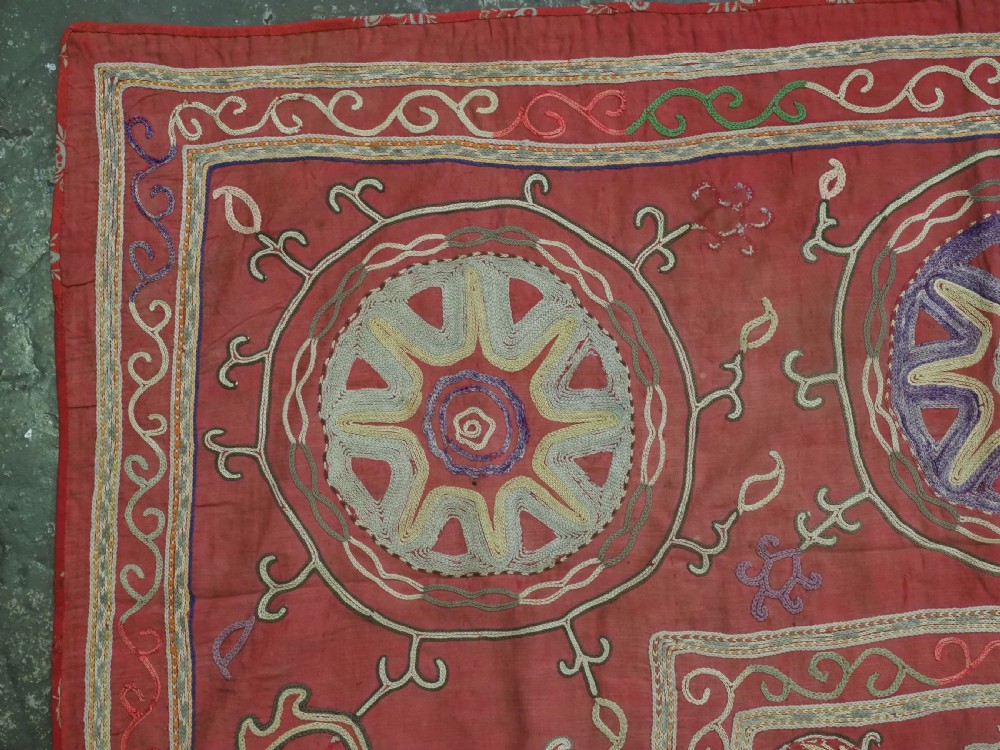 A rectangular red cotton embroidered suzani featuring geometric starburst medallions and floral scrolling borders worked in multi-coloured silk thread. View IV