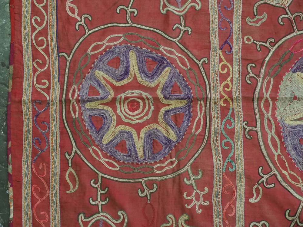 A rectangular red cotton embroidered suzani featuring geometric starburst medallions and floral scrolling borders worked in multi-coloured silk thread. View V