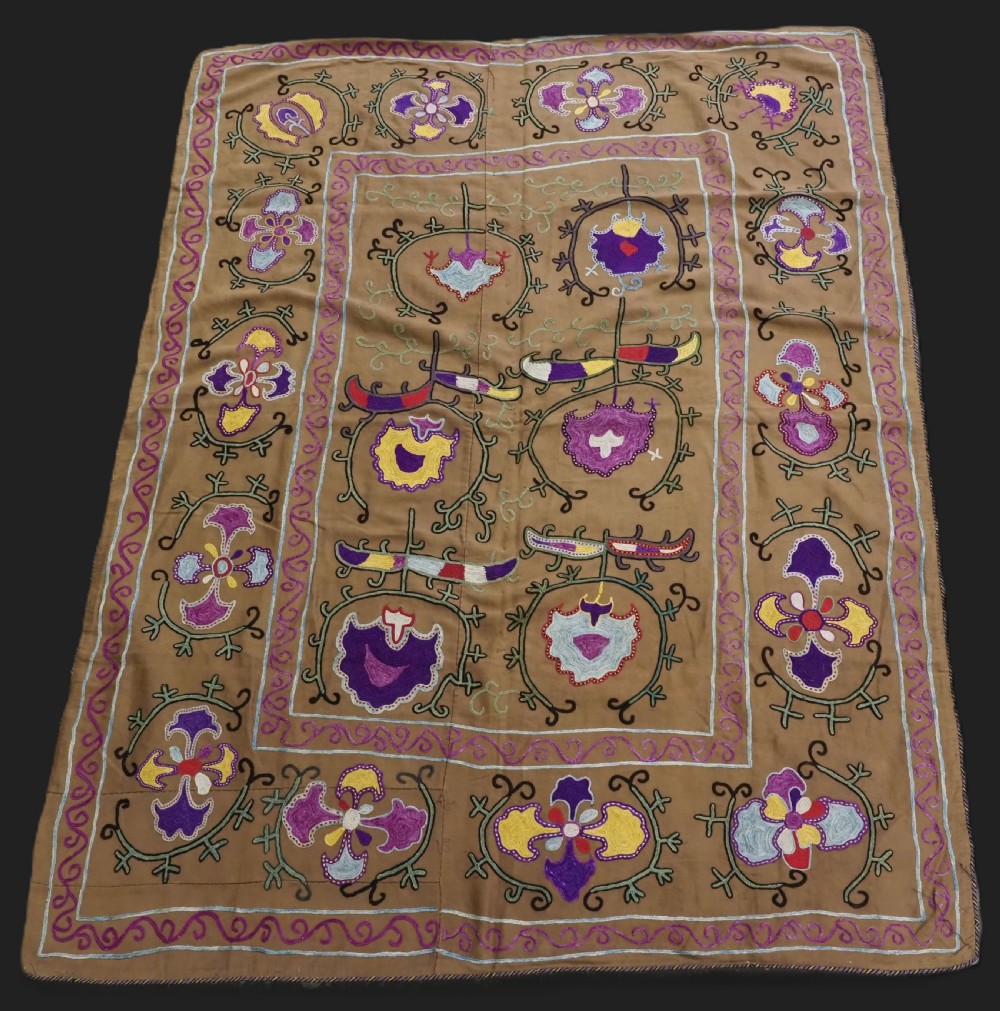 An antique Uzbek Nim Suzani textile featuring hand-embroidered floral motifs and hooked borders in purple, yellow, and blue silk threads on a tan cotton ground.