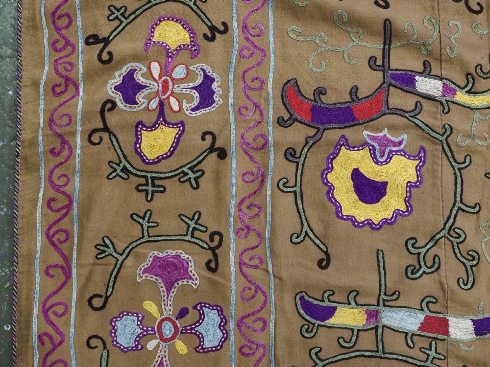 An antique Uzbek Nim Suzani textile featuring hand-embroidered floral motifs and hooked borders in purple, yellow, and blue silk threads on a tan cotton ground. View V