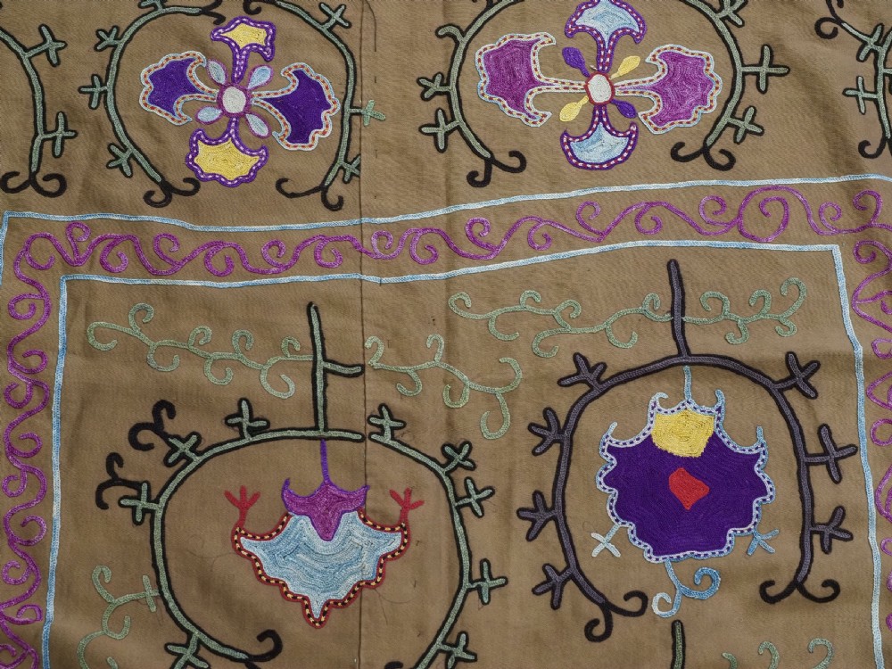 An antique Uzbek Nim Suzani textile featuring hand-embroidered floral motifs and hooked borders in purple, yellow, and blue silk threads on a tan cotton ground. View VI